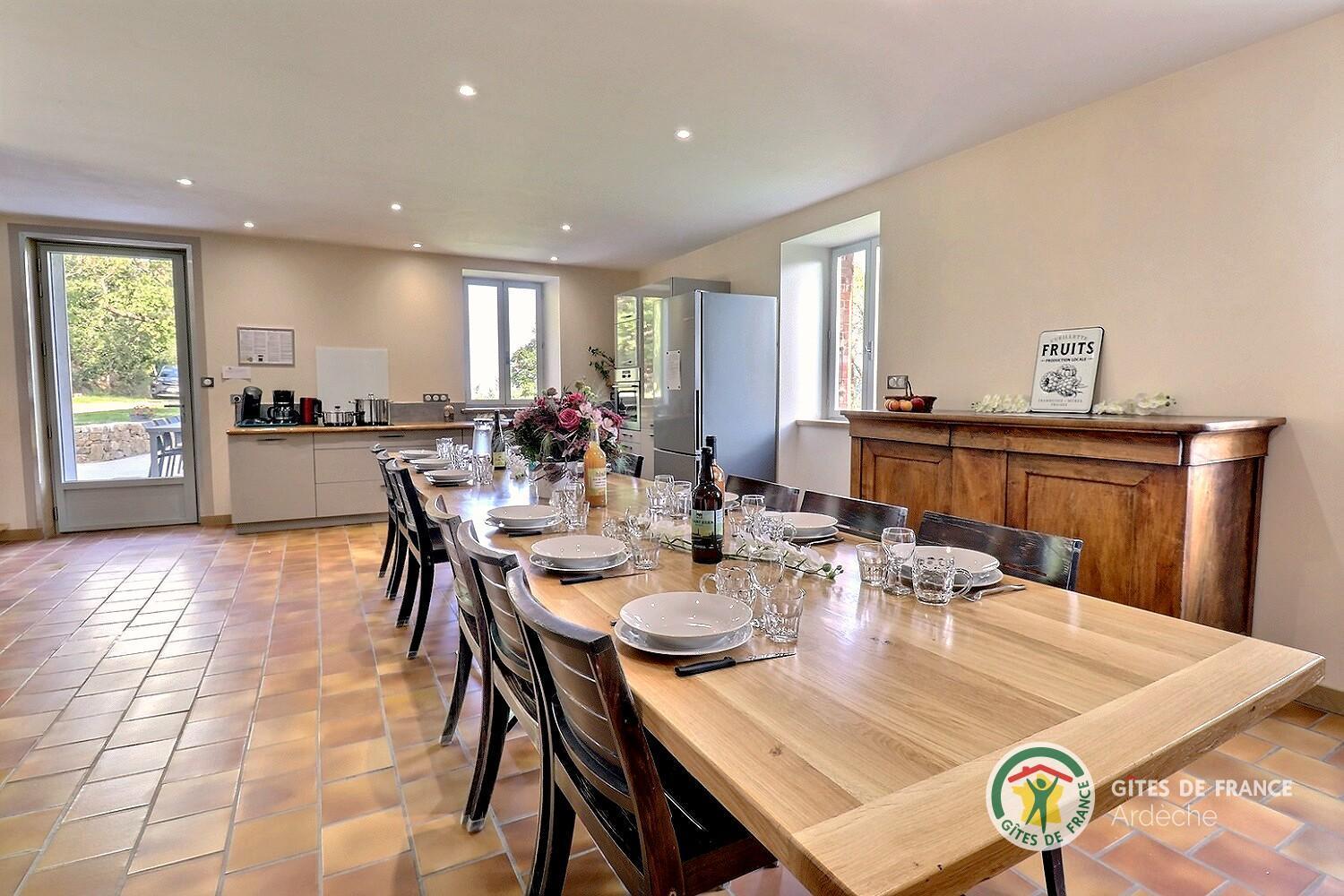 Grande salle à manger du gîte, longue table dressée pour dix couverts devant les fenêtres