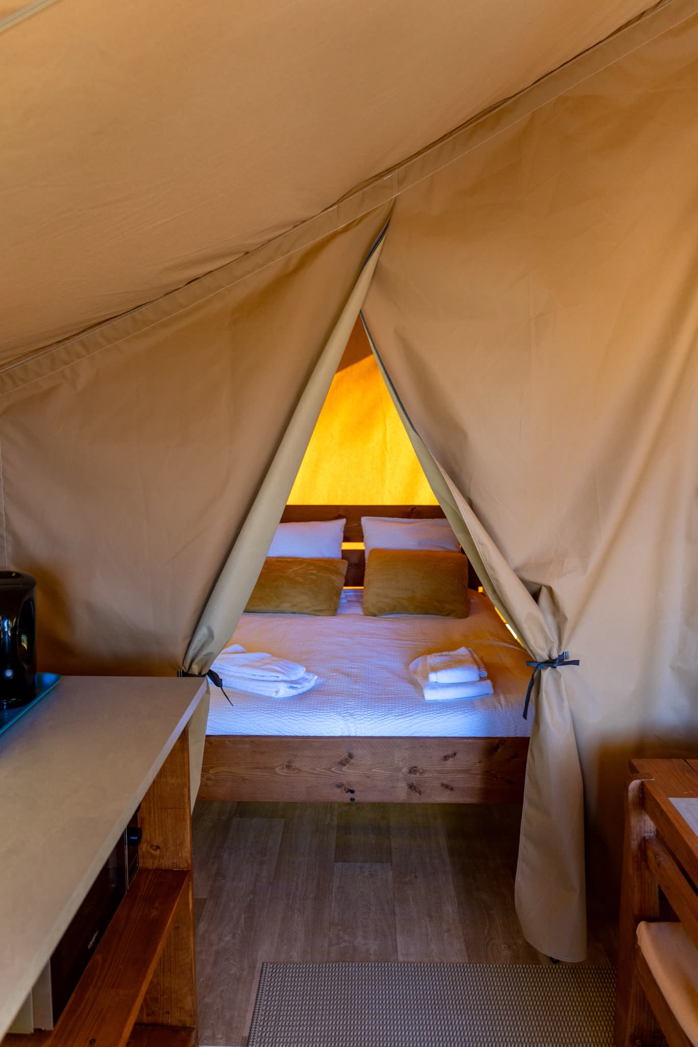 Chambre au lit double de la tente safari L'Estiva, tête de lit en bois, coussins ocre et linge de toilette préparé