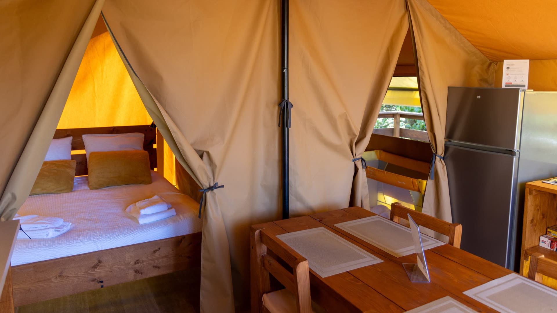 Intérieur de la tente safari L'Estiva : table en bois du coin repas, réfrigérateur et chambre au lit double derrière le rideau de toile