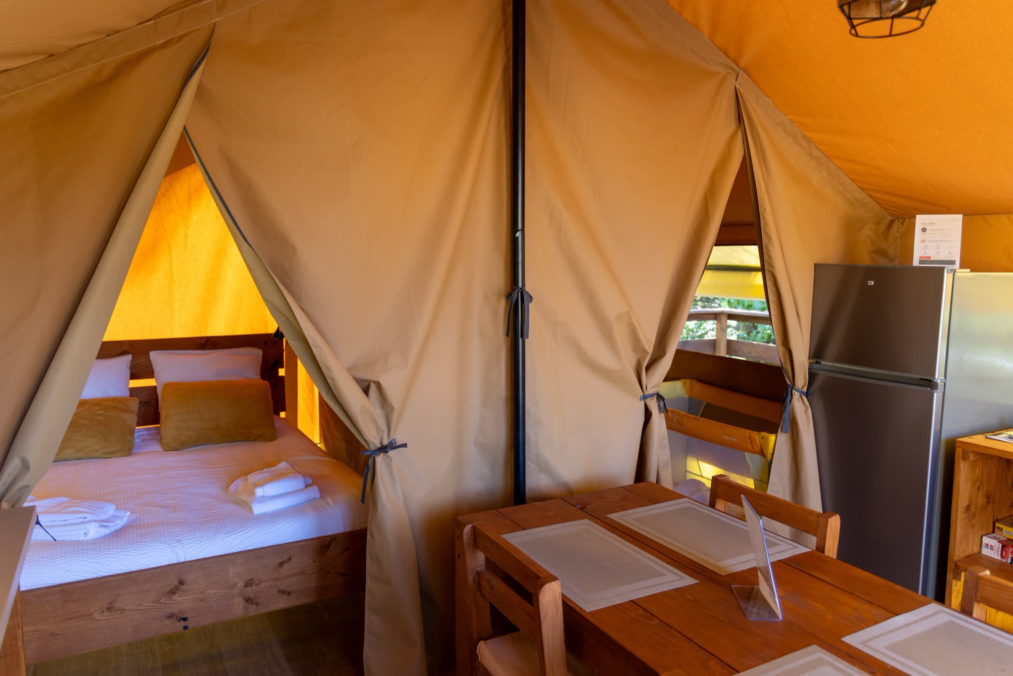 Intérieur de la tente safari L'Estiva : table en bois du coin repas, réfrigérateur et chambre au lit double derrière le rideau de toile