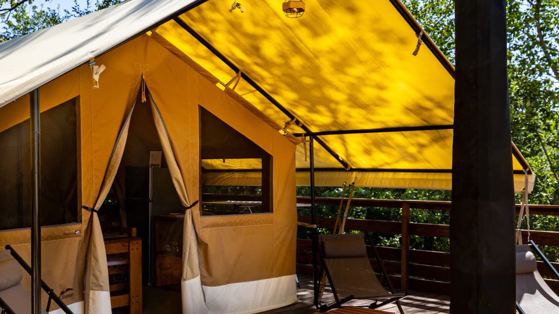 Terrasse en bois de 50 m² de la tente safari L'Estiva, auvent jaune relevé, transats et tabourets devant l'entrée en toile