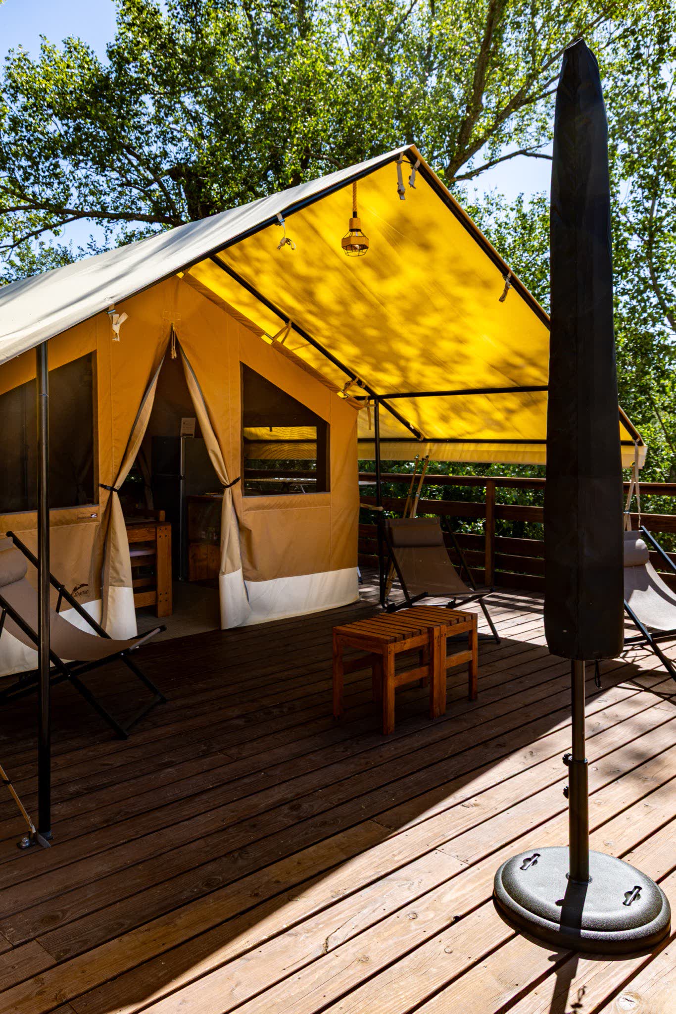 Terrasse en bois de 50 m² de la tente safari L'Estiva, auvent jaune relevé, transats et tabourets devant l'entrée en toile