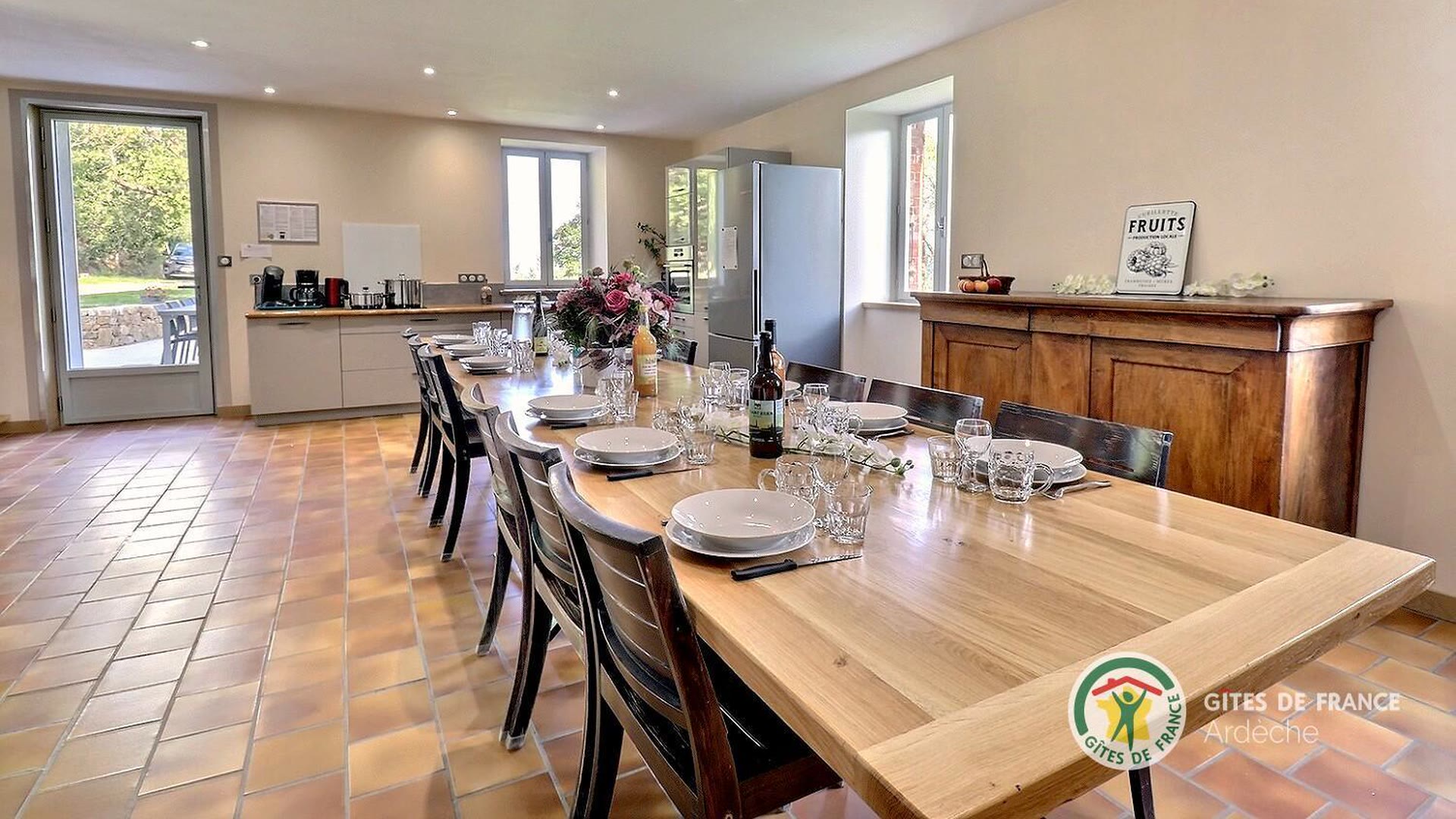 Grande salle à manger du gîte, longue table dressée pour dix couverts devant les fenêtres